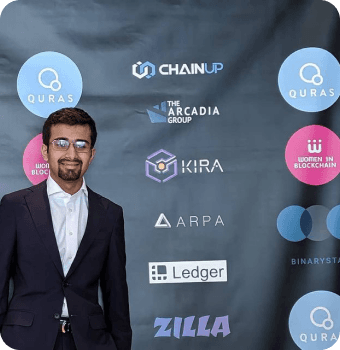 Rasikh Morani in a suit in front of a blockchain sponsor banner